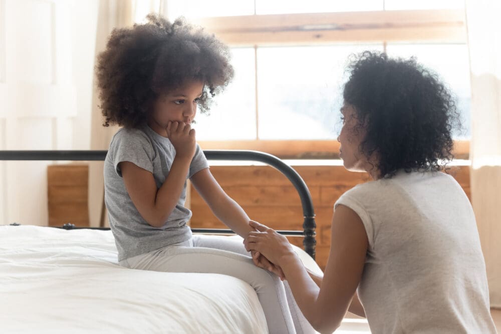 A concerned woman kneels by a bed, holding hands with a pensive young girl sitting on it
